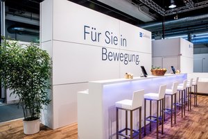 Modern gestalteter Messestand mit heller Theke, Hochstühlen und Werbeslogan