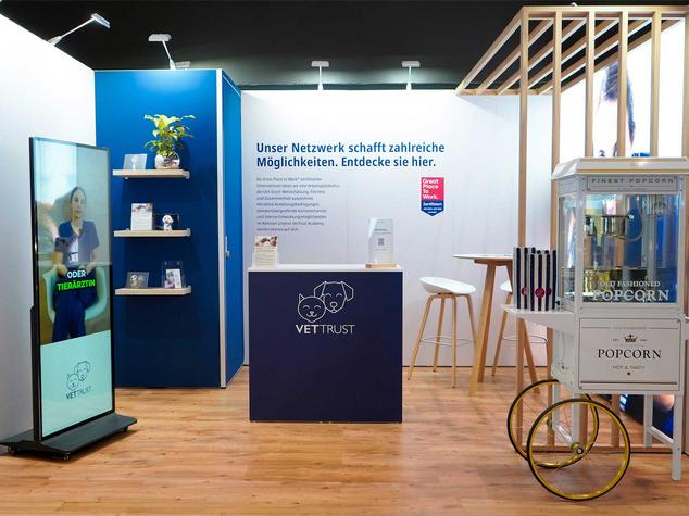 Ein Messestand mit interaktivem Bildschirm, Informationsregalen und einer Popcornmaschine.