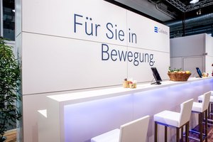 Professioneller Messestand mit Slogan 'Für Sie in Bewegung', Barhockern und einem Infoterminal