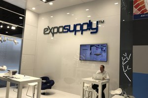 Moderner Messestand mit Marke Exposupply, beleuchtet, mit Ausstellungstischen und einer sitzenden Person am Laptop.