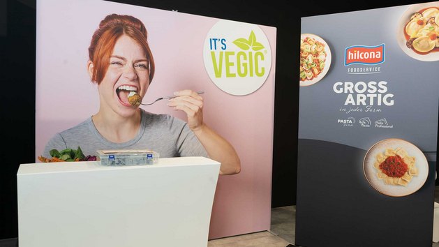 Eine Person isst mit einer Gabel und ein Werbebanner für pflanzliche Produkte ist im Hintergrund.