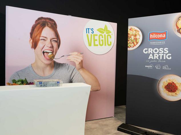 Eine Person isst mit einer Gabel und ein Werbebanner für pflanzliche Produkte ist im Hintergrund.