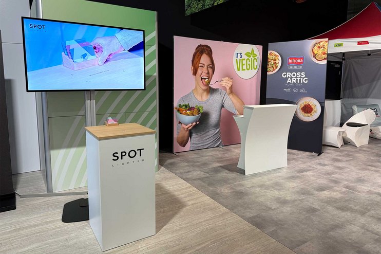 Ein Messestand mit Bildschirm, Theke und Werbebanner für vegane Produkte.