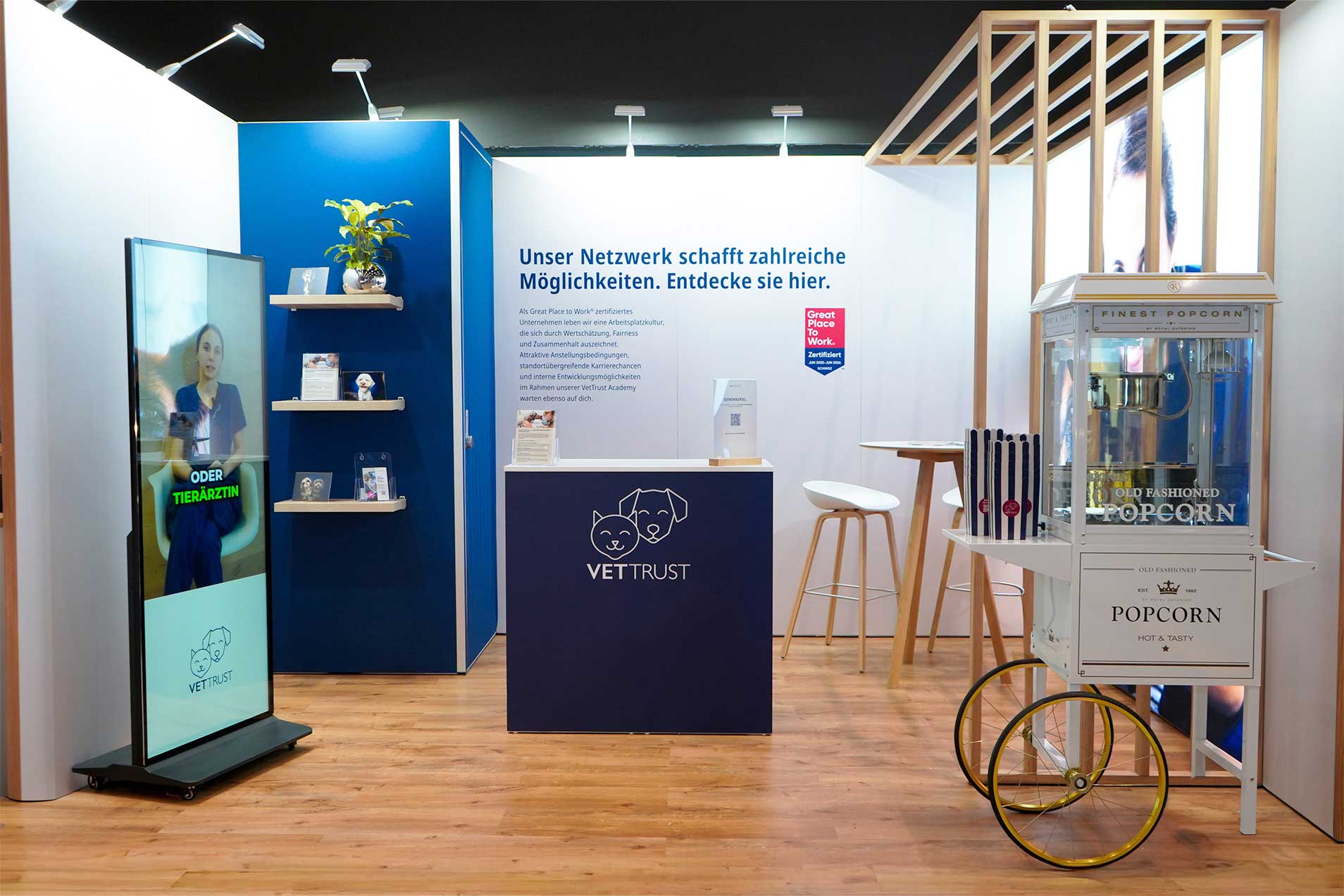 Ein Messestand mit interaktivem Bildschirm, Informationsregalen und einer Popcornmaschine.